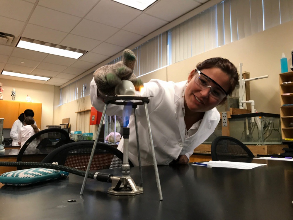 A scientist donning safety goggles and a lab coat uses a bunsen burner in a lab.